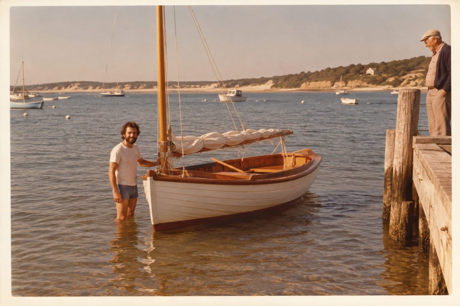 Tom standing in the water beside his newly launched catboat, an old man watching from the landing