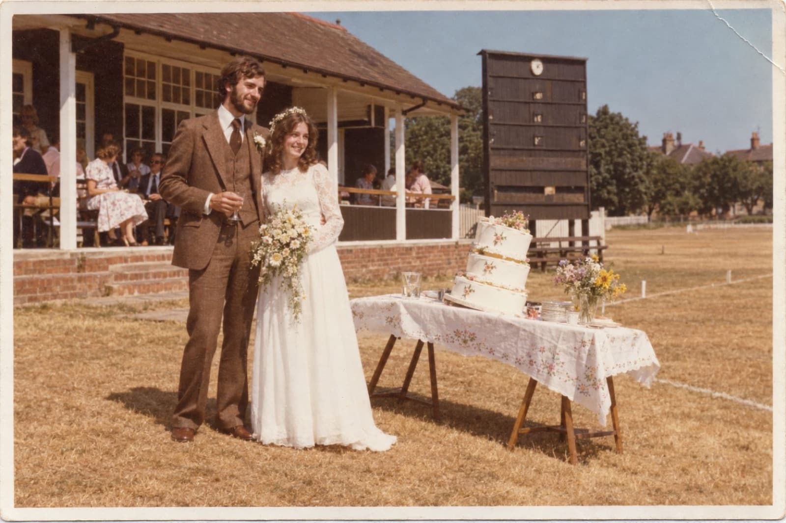 Richard and Anne on their wedding day in front of a cricket pavilion, the cake beside them leaning in the heat