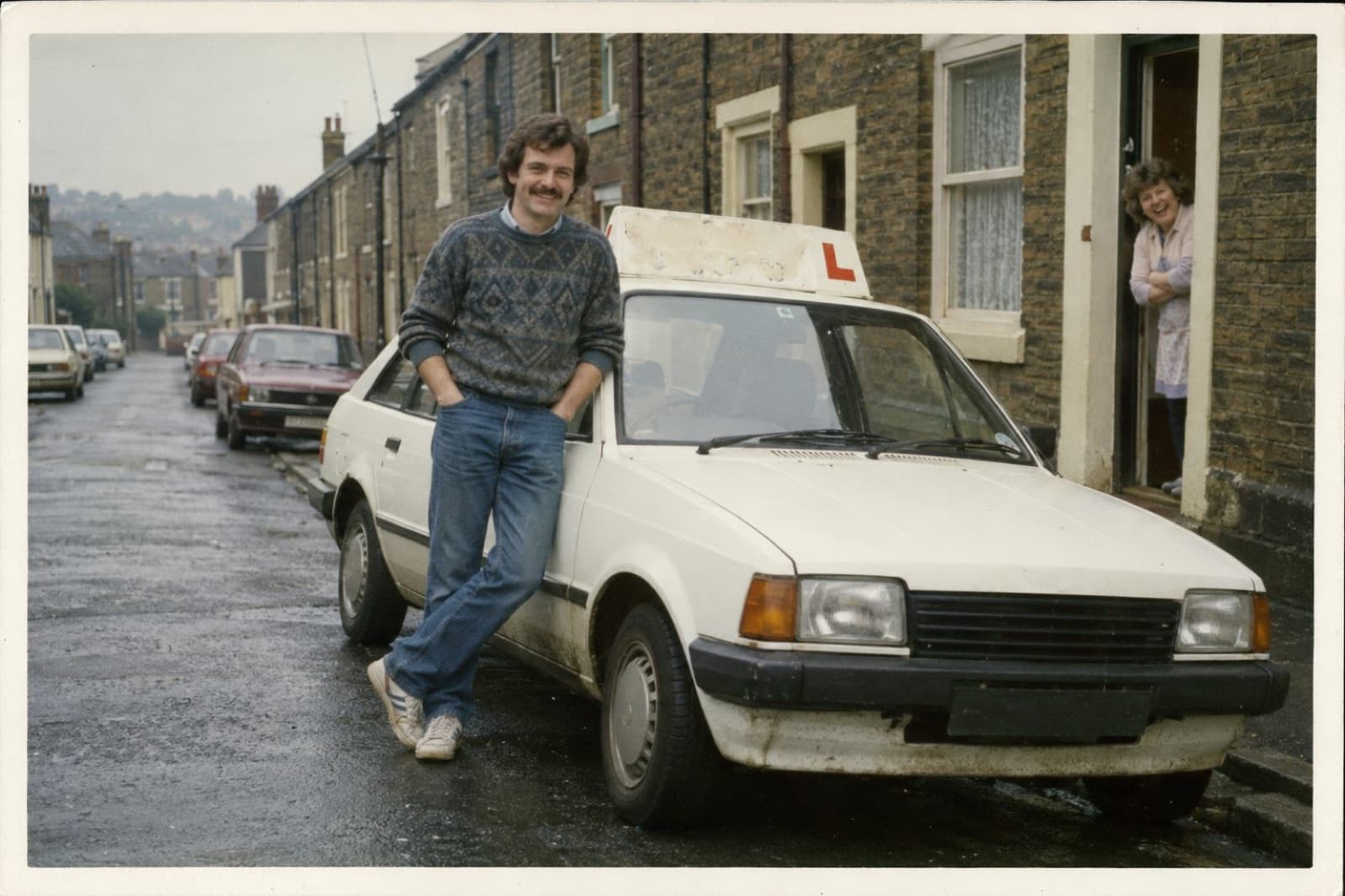 Graham leaning on a white Ford Escort with a sign on its roof, on a terraced street in Sheffield
