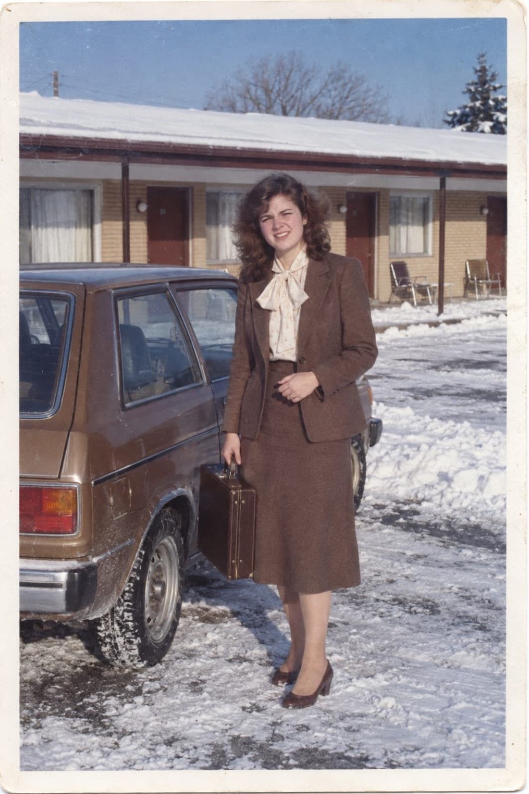 Carol in a brown skirt suit beside her car outside a motel in the snow